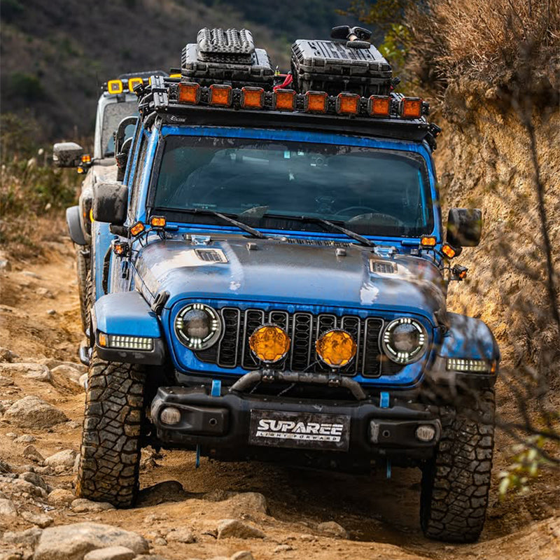 Suparee Helm 7 inch amber LED pods mounted on the front bumper and roof rack of a blue Jeep Wrangler driving off-road.